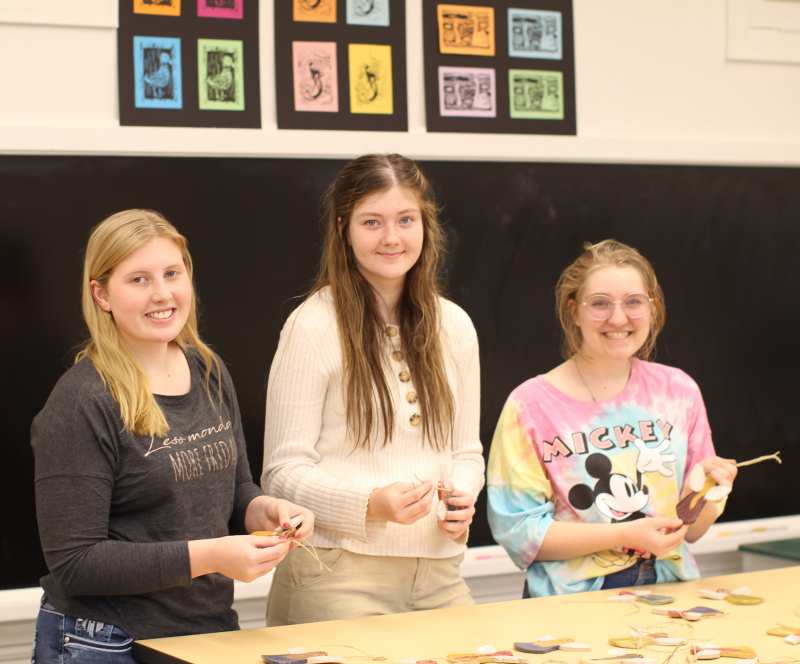 MC art education students, from left, Dani Henderson, Emily Pearson, and Cate Stennett prepare ornaments for the sale that will send senior art students to the NAEA Convention. Some of the print collages for sale are visible on the wall behind them.